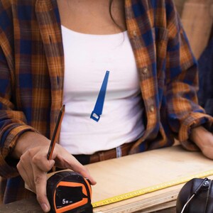 May include: A person using a tape measure and a pencil to measure a piece of wood. The tape measure is orange and black and reads "5m". The wood is light brown and has a yellow ruler along the edge.