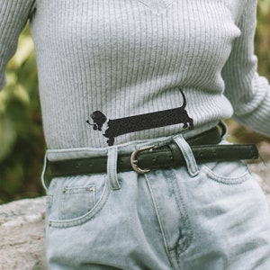 Könnte beinhalten: Ein grauer Pullover mit einem schwarzen Dachshund-Silhouetten-Design. Der Pullover wird über einer hellblauen Jeans mit einem schwarzen Gürtel getragen.