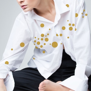 Könnte beinhalten: Ein weißes Hemd mit Knöpfen und goldenen Glitzerpunkten. Das Hemd wird von einer Person getragen, die mit gekreuzten Beinen sitzt.