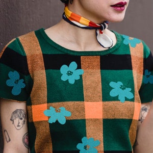 Könnte beinhalten: Ein grünes, orangefarbenes und schwarzes kariertes Hemd mit blauen Blumendesigns. Das Hemd hat einen runden Ausschnitt und kurze Ärmel.