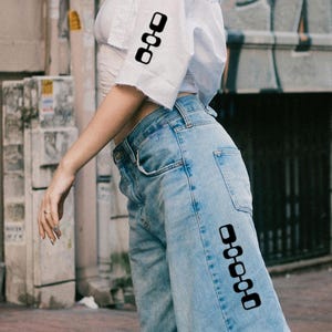 Könnte beinhalten: Eine Person trägt ein weißes Hemd mit schwarzen Quadraten am Ärmel und eine blaue Jeans mit schwarzen Quadraten am Bein.