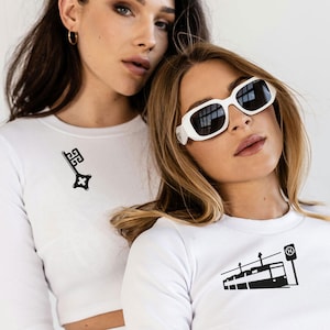 Puede incluir: Dos mujeres con camisetas blancas de manga larga con diseños gráficos negros. Una camiseta presenta un diseño de llave, la otra un tranvía. Una mujer lleva gafas de sol blancas. Las camisetas parecen ser acanaladas.