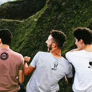 Puede incluir: Tres personas con camisetas con gráficos de temática cervecera. Una camiseta es rosa polvoriento con el logo "BREMEN", otra es gris con una copa de cerveza y la tercera es blanca con una jarra de cerveza. El fondo es un entorno natural verde.