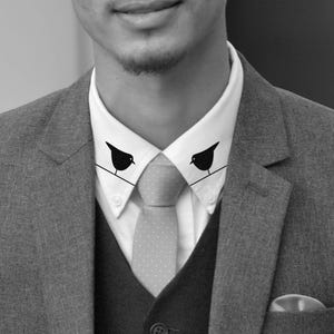 May include: A black and white photo of a person wearing a suit with a white collared shirt and a patterned tie. The collar of the shirt has a black bird silhouette on each side.