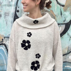 Op de afbeelding: Een witte trui met een hoge hals en zwarte bloemenborduursels. De trui heeft een losse pasvorm en een zachte, pluche textuur.