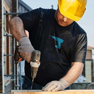 Op de afbeelding: Een bouwvakker met een gele helm en zwarte handschoenen gebruikt een boormachine om een ​​stuk hout aan een houten balk te bevestigen.
