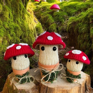Pode incluir: Cinco peluches de cogumelo de crochê em vários tamanhos, com chapéus vermelhos e brancos, sentados em um chão de floresta coberto de musgo. Os peluches têm olhos brancos e estão em vasos marrons com corda.