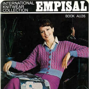 Può includere: Una donna indossa un cardigan di maglia rosa e blu con chiusura a bottoni. È seduta su una sedia e tiene una rivista. L'immagine è tratta dalla collezione Empisal International Knitwear, Libro AU26.