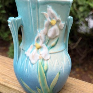 May include: A light blue ceramic vase featuring two side handles and a raised relief design of white iris flowers with yellow centers and green leaves. The surface has a subtle crackle glaze finish and a flared rim with vertical detailing.