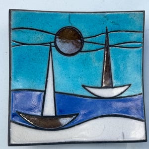 May include: A square ceramic dish featuring a hand-painted scene of two sailboats on a blue sea under a dark brown sun. The design uses bold black outlines, with white sails, a light blue sky, and a white foreground, finished with a glossy glaze.