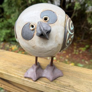 Op de afbeelding: Een rond keramieken vogelbeeldje met een crèmekleurig lichaam, donkergrijze oogvlekken en een puntige snavel. De vogel heeft donkergrijze zwemvliezen aan dunne metalen poten en decoratieve donkergrijze vleugelpanelen met lichtblauwe krulpatronen.