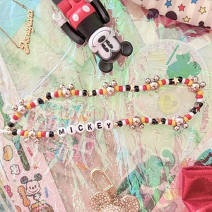 May include: A collection of Mickey Mouse themed items including a beaded necklace with the word "MICKEY" spelled out, a gold sparkly Mickey Mouse keychain, a red and black Mickey Mouse figurine, and a red and white Mickey Mouse fabric bag.