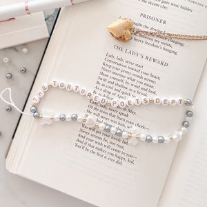May include: A white and silver beaded bracelet with a butterfly charm and the words "The Tortured Poets Dept" spelled out in white letters. The bracelet is lying on a book with the text "The Lady's Reward" and "Prisoner" visible.
