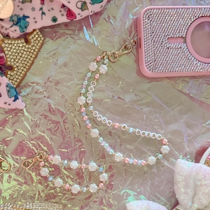 Puede incluir: Un charm de teléfono de cuentas rosa y blanco con las palabras "Once Upon A Dream" escritas en cuentas blancas. El charm está unido a una cadena dorada y está rodeado de otros accesorios de temática de princesa, incluyendo una funda de teléfono rosa con pedrería, una diadema rosa con pedrería y un lazo rosa y blanco.