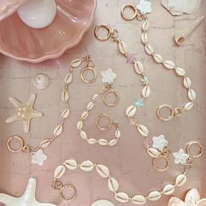 Puede incluir: Colección de llaveros y accesorios de conchas marinas. Los llaveros presentan conchas marinas naturales unidas con detalles y cierres metálicos dorados. Algunos llaveros incluyen dijes de flores y estrellas blancas. El fondo incluye conchas marinas y un plato rosa en forma de concha.