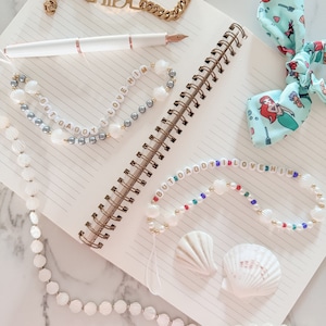 May include: A notebook with a gold chain bracelet, a white pen, and three shell necklaces with the words "But Daddy I Love Him" on them. There is also a blue and green hair bow with a mermaid design on it.
