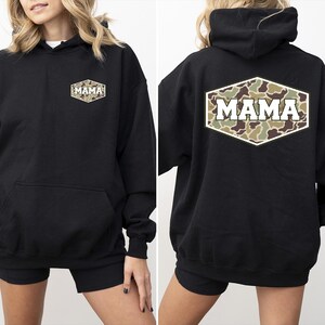 May include: Black hooded sweatshirt with a front pocket and the word "MAMA" in white block letters on the back. The word "MAMA" is inside a camouflage-patterned, hexagonal shape. A smaller version of the same design is on the front left chest.