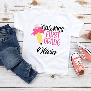 May include: White t-shirt with a pink and yellow pencil graphic and the text "Little Miss First Grade Olivia".