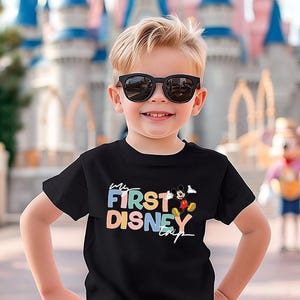 May include: A young child wearing a black t-shirt with the text "My First Disney Trip" in colorful letters. The shirt also features a graphic of Mickey Mouse. The child is smiling and wearing sunglasses, with a Disney castle in the background.