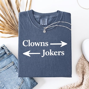 May include: A navy blue t-shirt with the words "Clowns" and "Jokers" separated by an arrow. A silver necklace with a small pendant rests on the shirt. The shirt is folded with a pair of blue jeans and white slip-on shoes nearby.