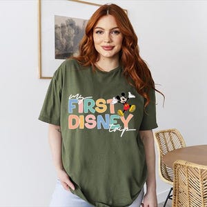 May include: Olive green t-shirt with the text "My First Disney Trip" in colorful letters and a graphic of Mickey Mouse. The shirt is a relaxed fit and has short sleeves.