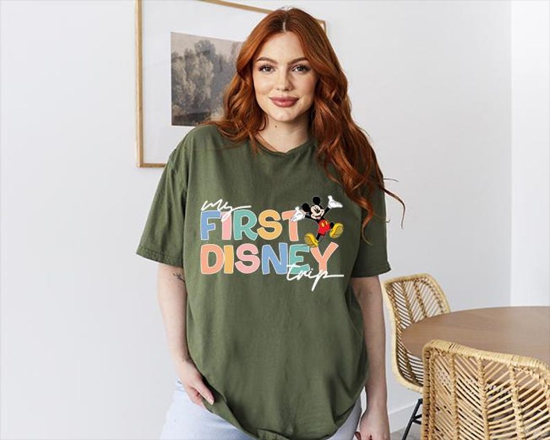 May include: Olive green t-shirt with the text "My First Disney Trip" in colorful letters and a graphic of Mickey Mouse. The shirt is a relaxed fit and has short sleeves.