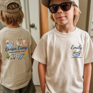 Pu&ograve; includere: Una t-shirt beige con stampa grafica. La parte anteriore mostra il nome Emily sopra una piccola illustrazione di una piscina e il testo Cousin Camp 2026. Il retro presenta una grafica pi&ugrave; grande con il testo Cousin Camp Mama and Papa 2026.