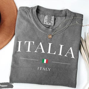 May include: A gray Comfort Colors t-shirt with the word "ITALIA" in white block letters. Below the word is a horizontal line with the Italian flag and the word "ITALY". A silver necklace with a pendant rests on the shirt.