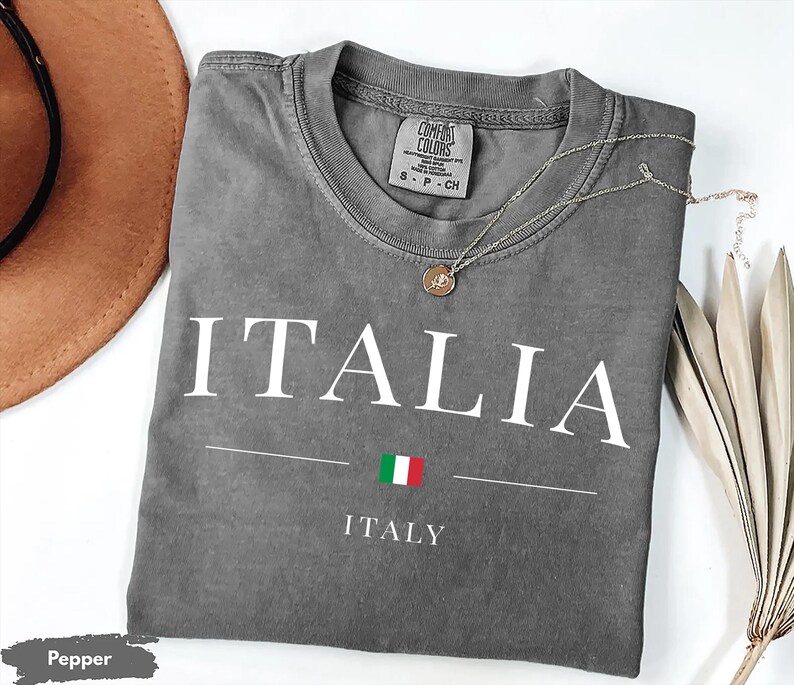 May include: A gray Comfort Colors t-shirt with the word "ITALIA" in white block letters. Below the word is a horizontal line with the Italian flag and the word "ITALY". A silver necklace with a pendant rests on the shirt.