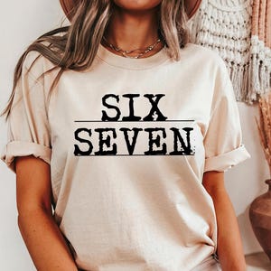 May include: A beige t-shirt with the words "SIX" over "SEVEN" in bold, black letters. The text is centered and underlined with a black line. The shirt has short sleeves and a relaxed fit.