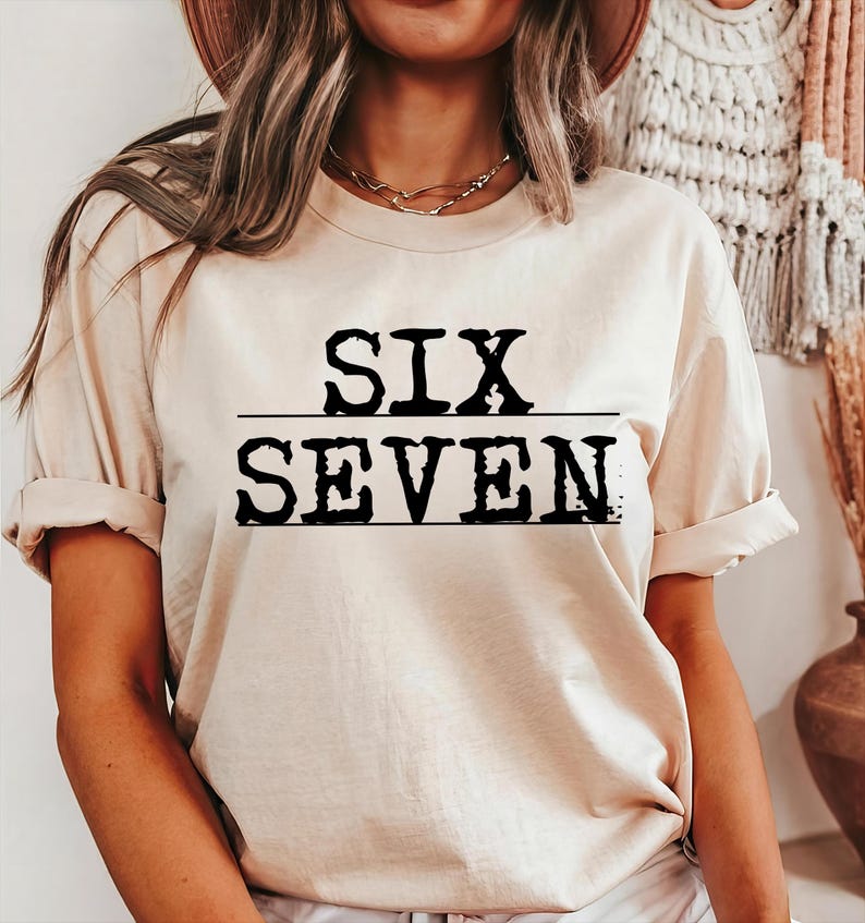 May include: A beige t-shirt with the words "SIX" over "SEVEN" in bold, black letters. The text is centered and underlined with a black line. The shirt has short sleeves and a relaxed fit.
