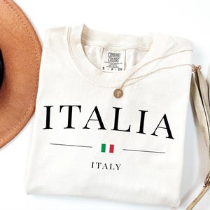May include: Cream-colored t-shirt with the word "ITALIA" in large black letters, a horizontal line, and the Italian flag. Below the line is the word "ITALY". A gold necklace and a brown hat are also in the image.