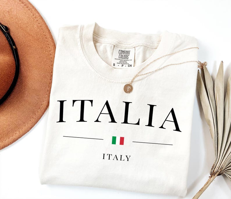 May include: Cream-colored t-shirt with the word "ITALIA" in large black letters, a horizontal line, and the Italian flag. Below the line is the word "ITALY". A gold necklace and a brown hat are also in the image.