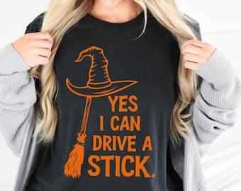 May include: Black t-shirt with an orange witch hat and broom graphic. The text reads "YES I CAN DRIVE A STICK." The design is a Halloween-themed graphic.