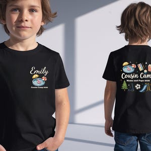 Pu&ograve; includere: Una t-shirt nera con una piccola grafica sul petto sinistro con il nome Emily, una piscina e il testo Cousin Camp 2026. Il retro mostra una grafica pi&ugrave; grande con il testo Cousin Camp Mama and Papa 2026 circondato da varie icone.
