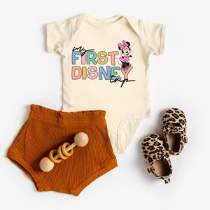 May include: A cream-colored baby onesie with the text "My First Disney Trip" and a Minnie Mouse graphic, paired with rust-colored shorts, a wooden rattle, and leopard print baby shoes.