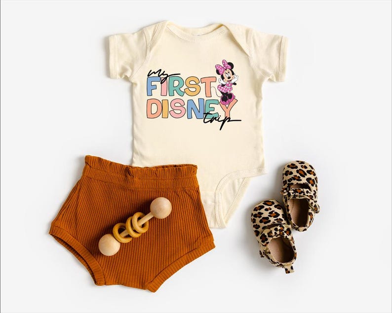 May include: A cream-colored baby onesie with the text "My First Disney Trip" and a Minnie Mouse graphic, paired with rust-colored shorts, a wooden rattle, and leopard print baby shoes.