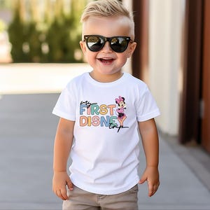May include: A white t-shirt with the text "My First Disney Trip" in colorful letters and a Minnie Mouse graphic. The shirt is worn by a young child wearing black sunglasses.