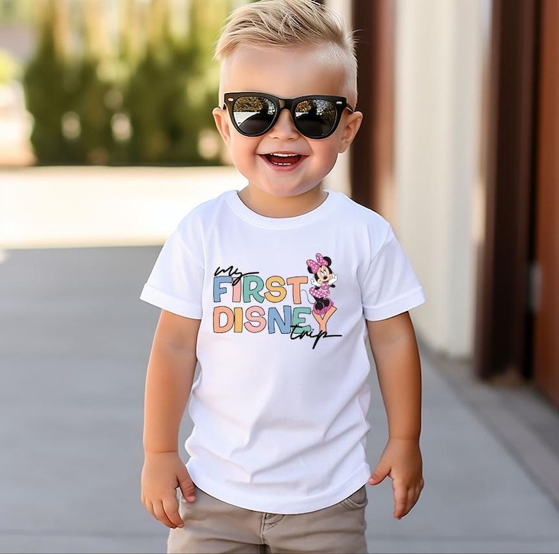 May include: A white t-shirt with the text "My First Disney Trip" in colorful letters and a Minnie Mouse graphic. The shirt is worn by a young child wearing black sunglasses.