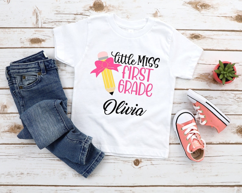 May include: White t-shirt with a pink and yellow pencil graphic and the text "Little Miss First Grade Olivia".