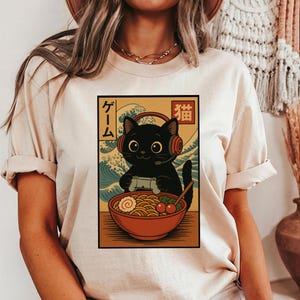 May include: Beige t-shirt featuring a black cat wearing a headset and holding a game controller. The cat is in front of a bowl of ramen. Japanese text is present. The background has a wave pattern.