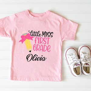May include: Pink t-shirt with a yellow pencil with a pink bow and the text "little miss first grade Olivia" on it.