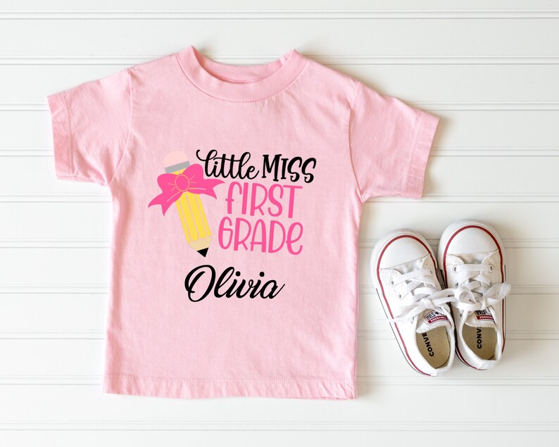 May include: Pink t-shirt with a yellow pencil with a pink bow and the text "little miss first grade Olivia" on it.