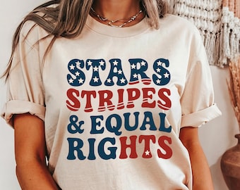 May include: A beige t-shirt with the words "STARS STRIPES & EQUAL RIGHTS" in blue and red lettering. The word "STARS" has white stars inside the blue letters. The shirt has rolled-up sleeves.