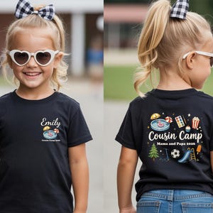 Pu&ograve; includere: Una t-shirt nera a maniche corte con una piccola grafica sul petto che riporta il nome Emily e una piscina. Il retro presenta una grafica pi&ugrave; grande con il testo Cousin Camp Mama and Papa 2026, circondato da icone di piscina, pallone e albero.