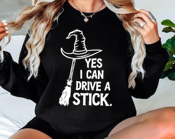 May include: Black sweatshirt with a white graphic of a witch's hat and broom, with the text "YES I CAN DRIVE A STICK." The sweatshirt is a crew neck style.