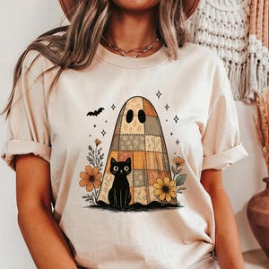 May include: Beige t-shirt featuring a Halloween-themed graphic. The design includes a patchwork ghost, a black cat, flowers, and a bat. The shirt has short sleeves and a relaxed fit, perfect for casual wear during the spooky season.