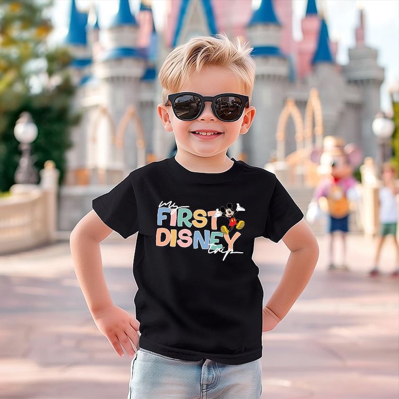 May include: A young child wearing a black t-shirt with the text "My First Disney Trip" in colorful letters. The shirt also features a graphic of Mickey Mouse. The child is smiling and wearing sunglasses, with a Disney castle in the background.