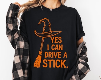 May include: Black t-shirt with an orange witch hat and broom graphic. The text "YES I CAN DRIVE A STICK" is printed below the graphic. The shirt is a Halloween-themed design.