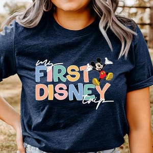 May include: Navy blue t-shirt with the text "My First Disney Trip" in colorful letters and a graphic of Mickey Mouse. The shirt is a casual style, suitable for a Disney vacation or themed event.